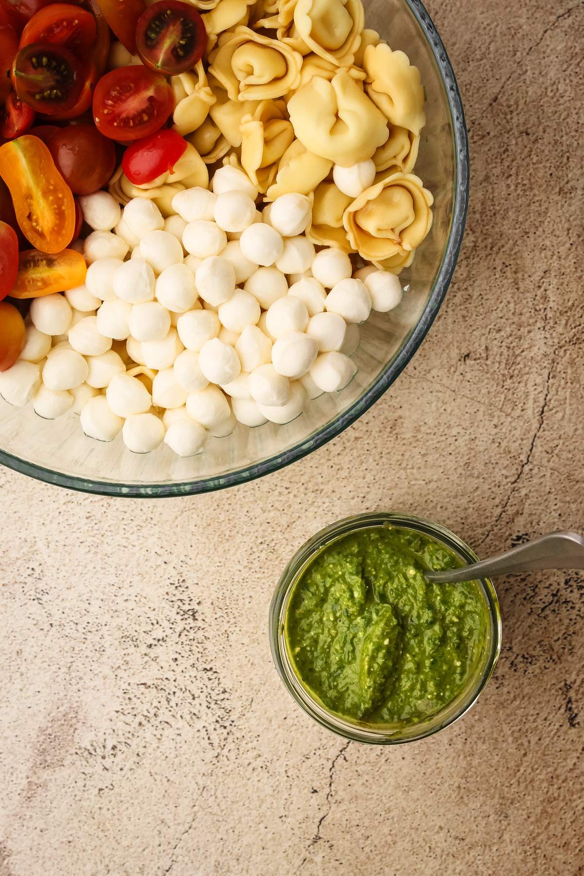 The ingredients for the tortollini salad each individually lined up in a bowl next to a container of pesto.