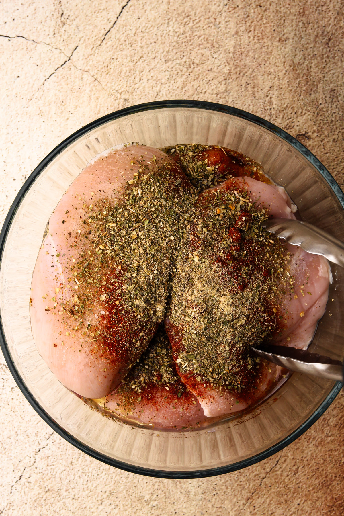 Raw chicken breasts in a bowl with spices on top before being mixed together.