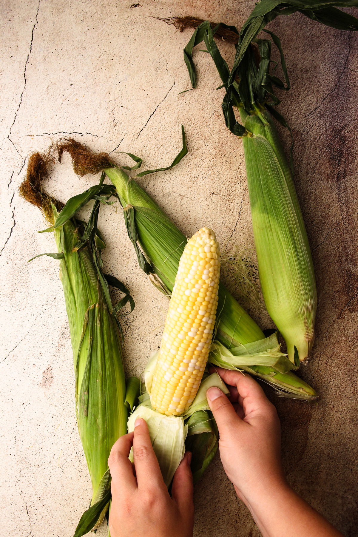 4 ears of corn with one getting shucked on a brown background.