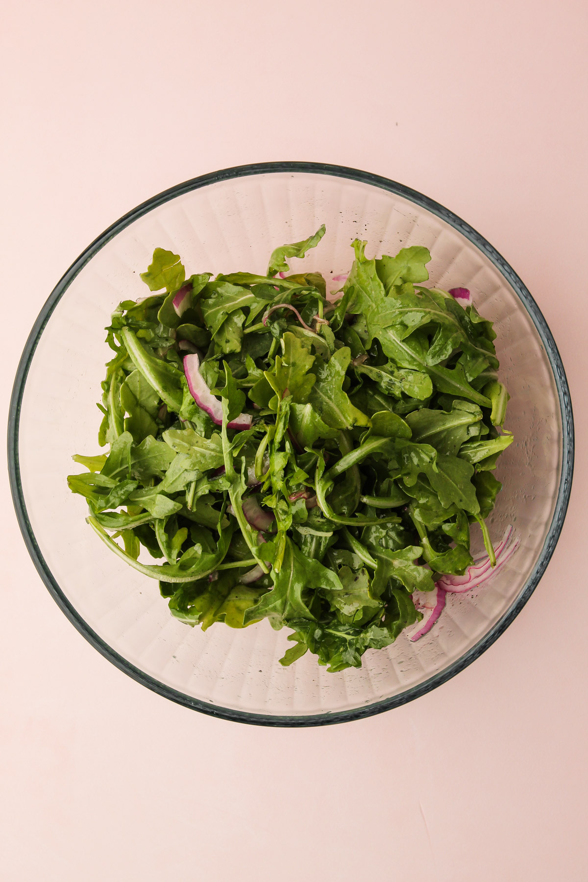 Arugula in a bowl with the dressing for a peach avocado salad on a pink background.