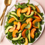 A platter with peach avocado salad with leaves of basil and arugula on a pink background.