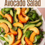 Peach and avocado salad with greens on a white plate with a pink background.