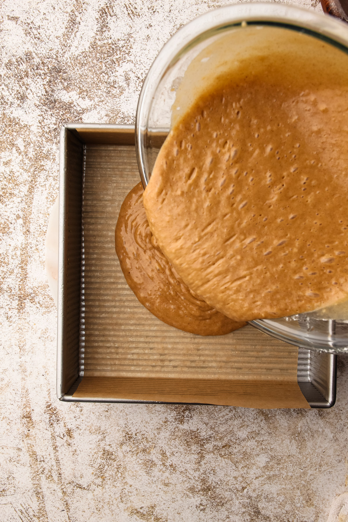 A brown, bubbly cake batter being poured from a glass bowl into a parchment-lined 8-inch square baking pan