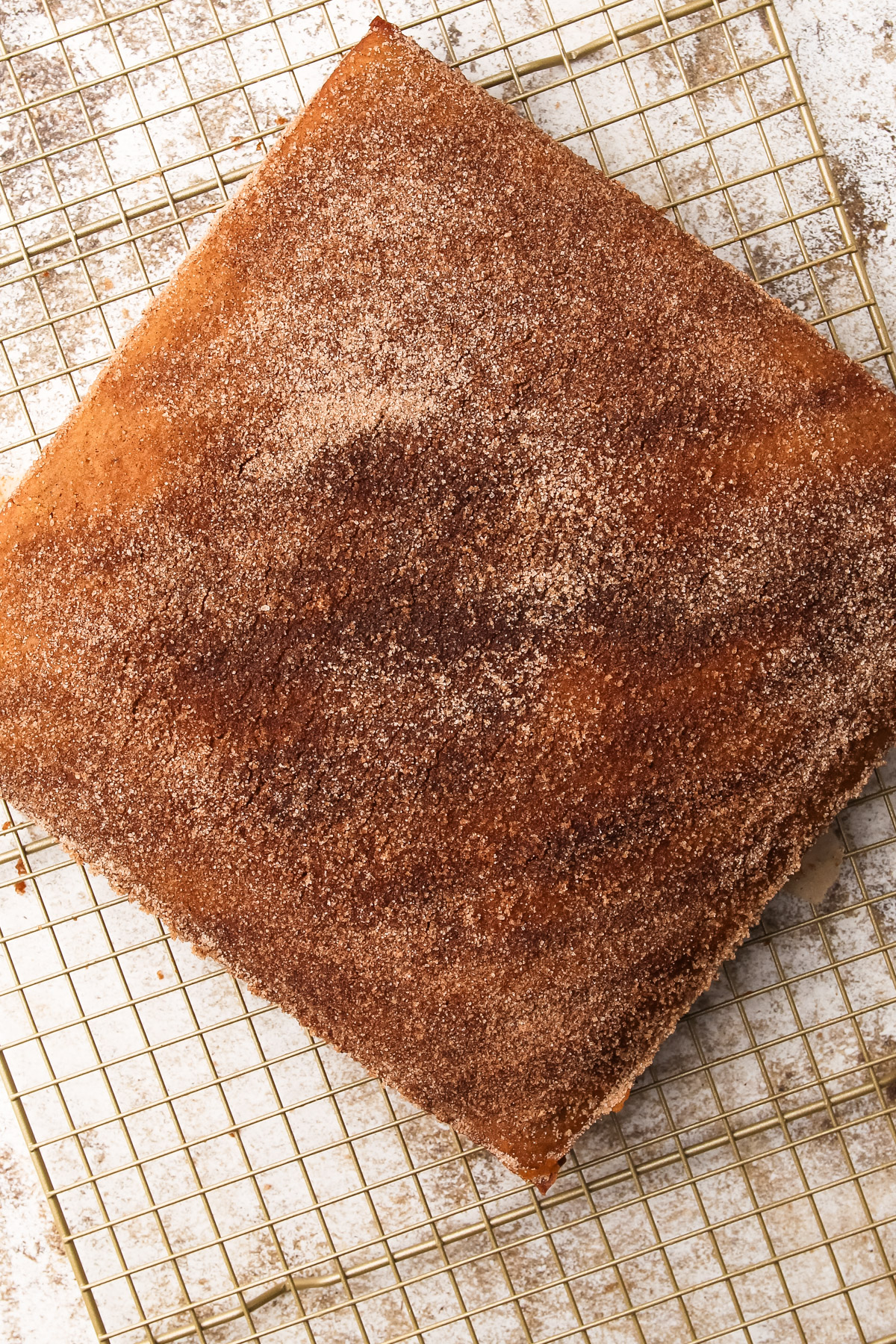 A square apple cideer donut cake on a cooling rack, sprinkled all over with cinnamon sugar on top.