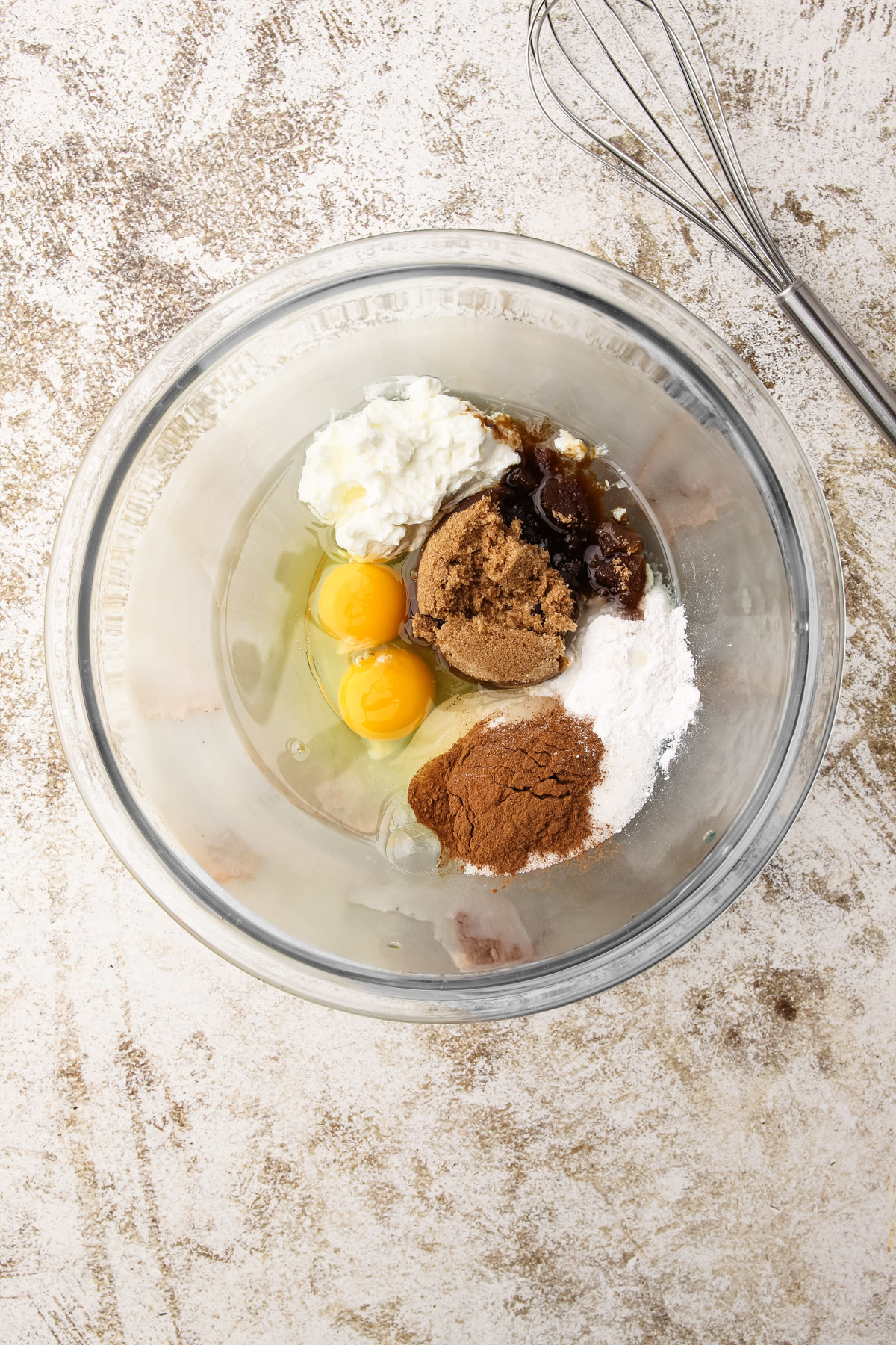 A glass bowl with flour, white sugar, brown sugar, cinnamon, eggs, and oil in it, with a whisk next to the bowl on the counter.