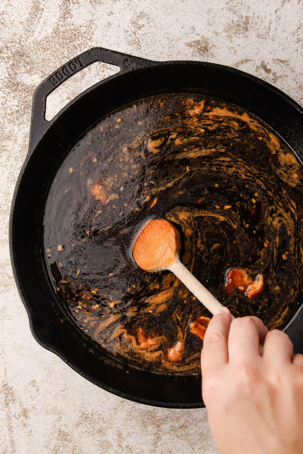 A hand using a wooden spoon to scrape the bottom of a cast iron skillet that has sauce and fond in it