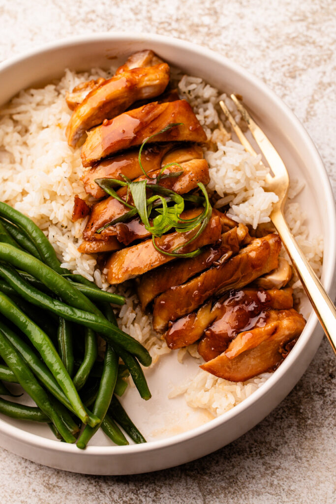 Slices of chicken thighs with teriyaki sauce drizzled over the top and topped with sliced scallions, on top of a bed of rice and next to a pile of cooked green beans, all in a deep plate with a fork.