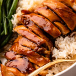 Slices of cooked chicken thighs with teriyaki sauce drizzled over the top, on top of a bed of rice and next to a pile of cooked green beans, all in a deep plate with a fork.