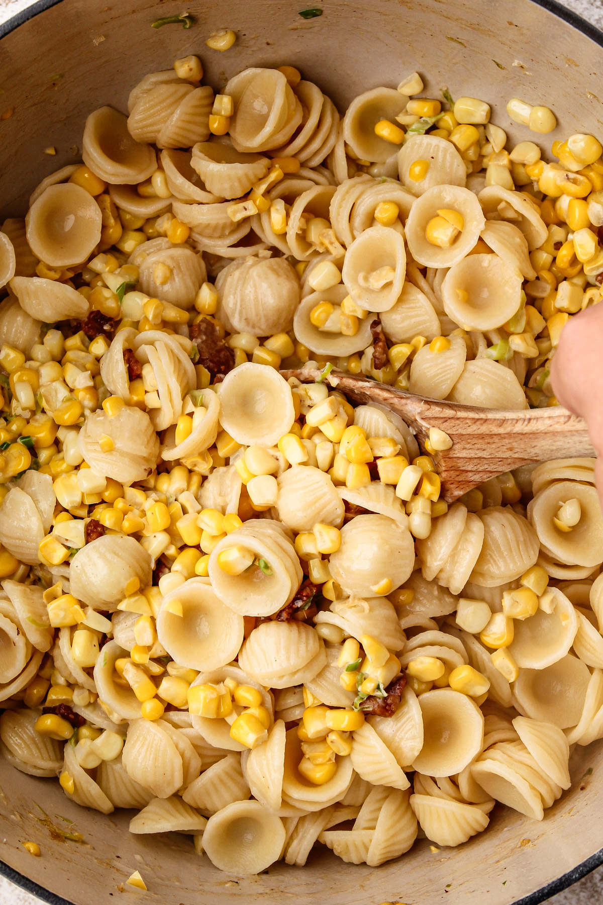 Stirring together pasta into the corn in bacon in a white dutch oven.