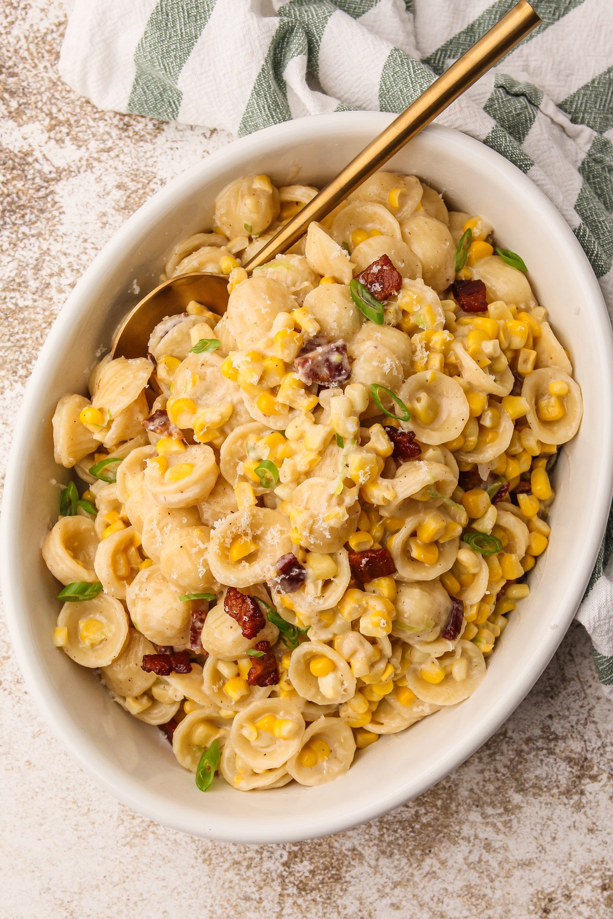 The finished corn pasta dish with bacon in a white serving dish next to a stripped green towel.