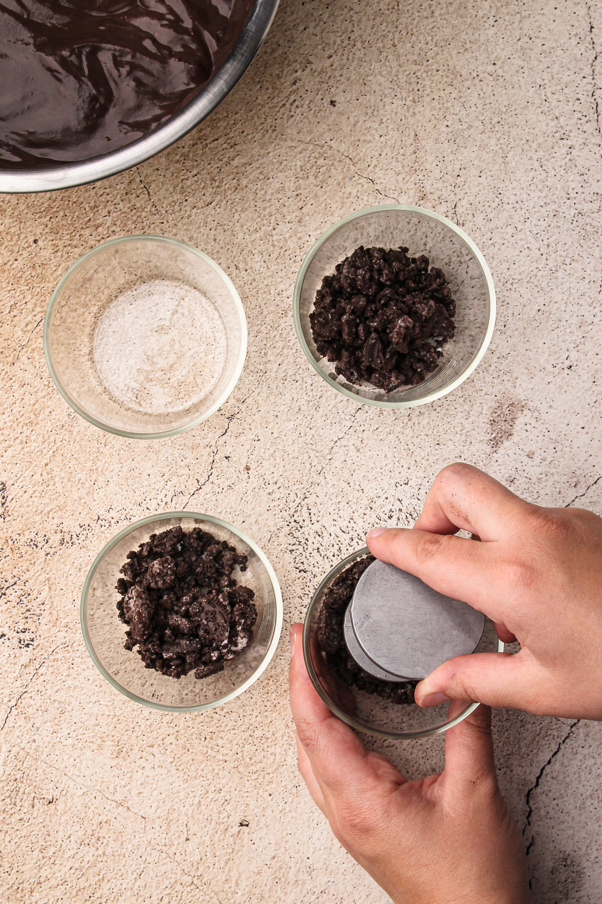 A hand using a tool to press crushed chocolate cookies into the bottom of a glass for Dirt Pudding Cups, with 3 more glasses nearby.