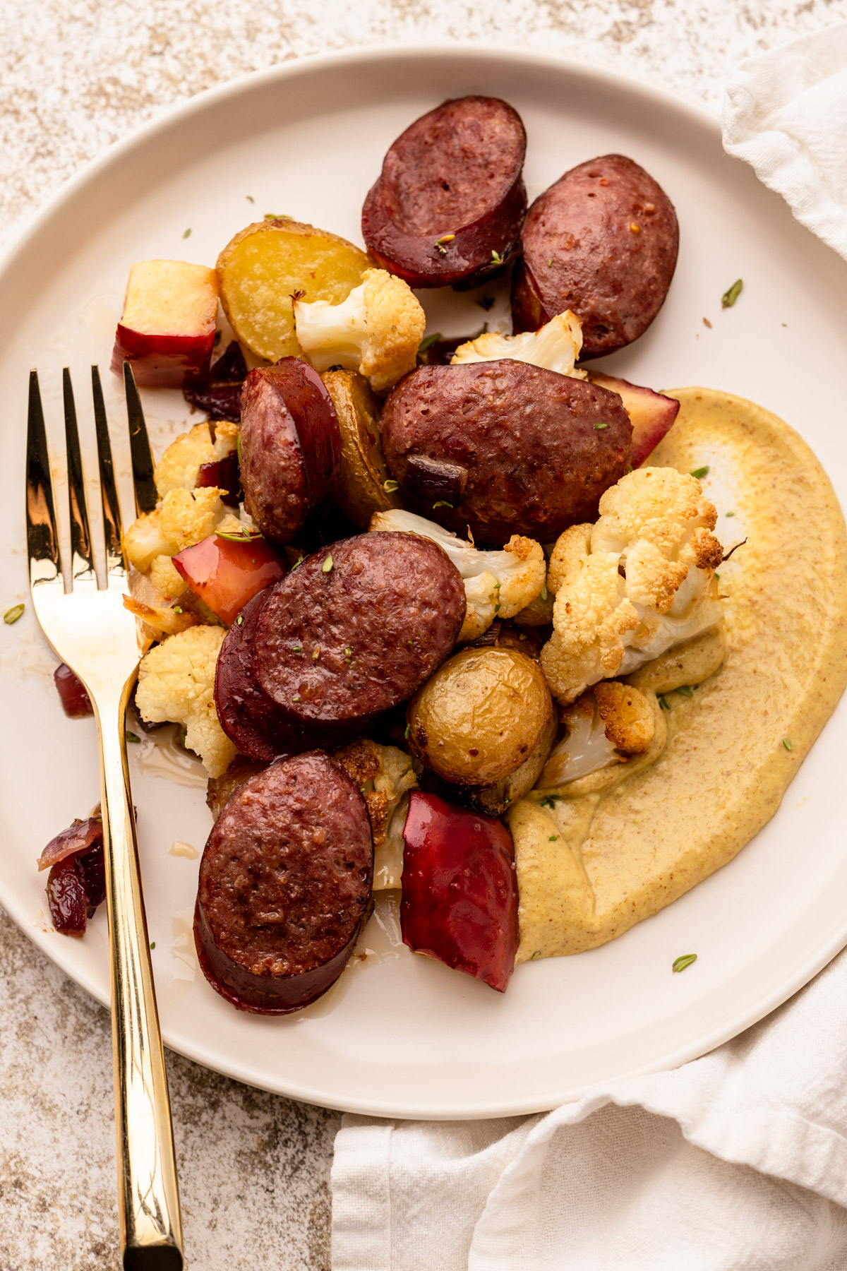 Roasted sheetpan kielbasa with apples, vegetables, and a swoosh of mustard sauce on a plate with a fork.