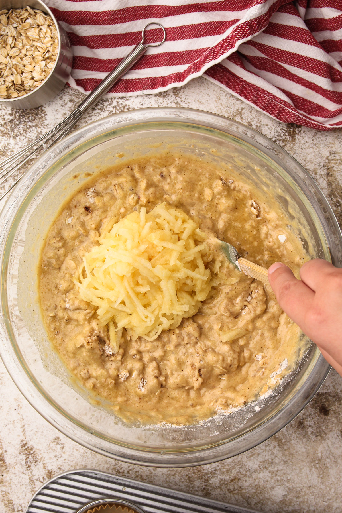 A hand using a rubber spatula to stir in shredded apple to a bowl of wholesome apple muffin batter.