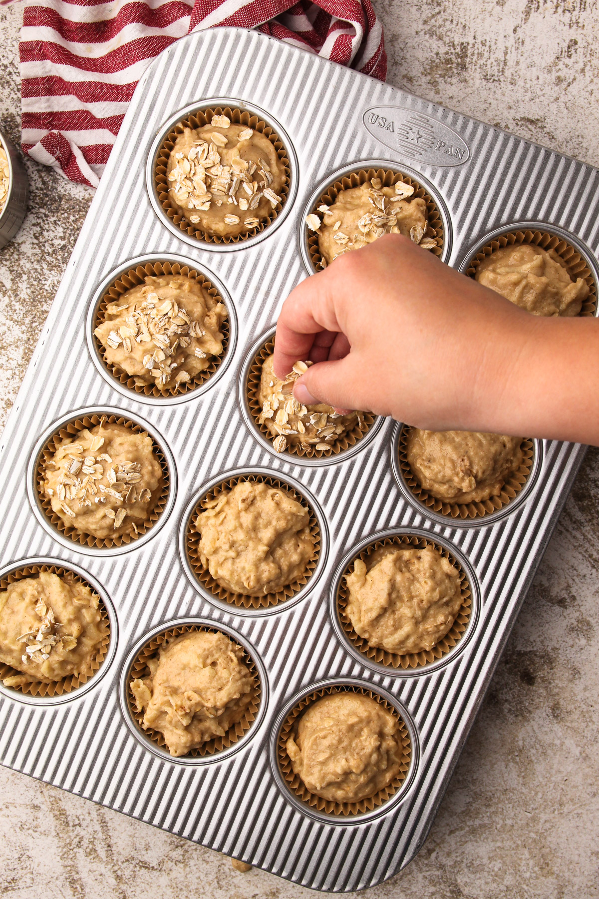 A hand sprinkling oats onto portions of wholesome apple muffin batter in liners in a metal muffin tin.