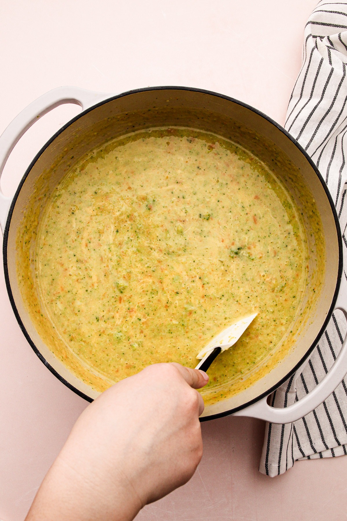 A hand using a rubber spatula to stir Broccoli Cheddar Bacon Soup in a Dutch oven.
