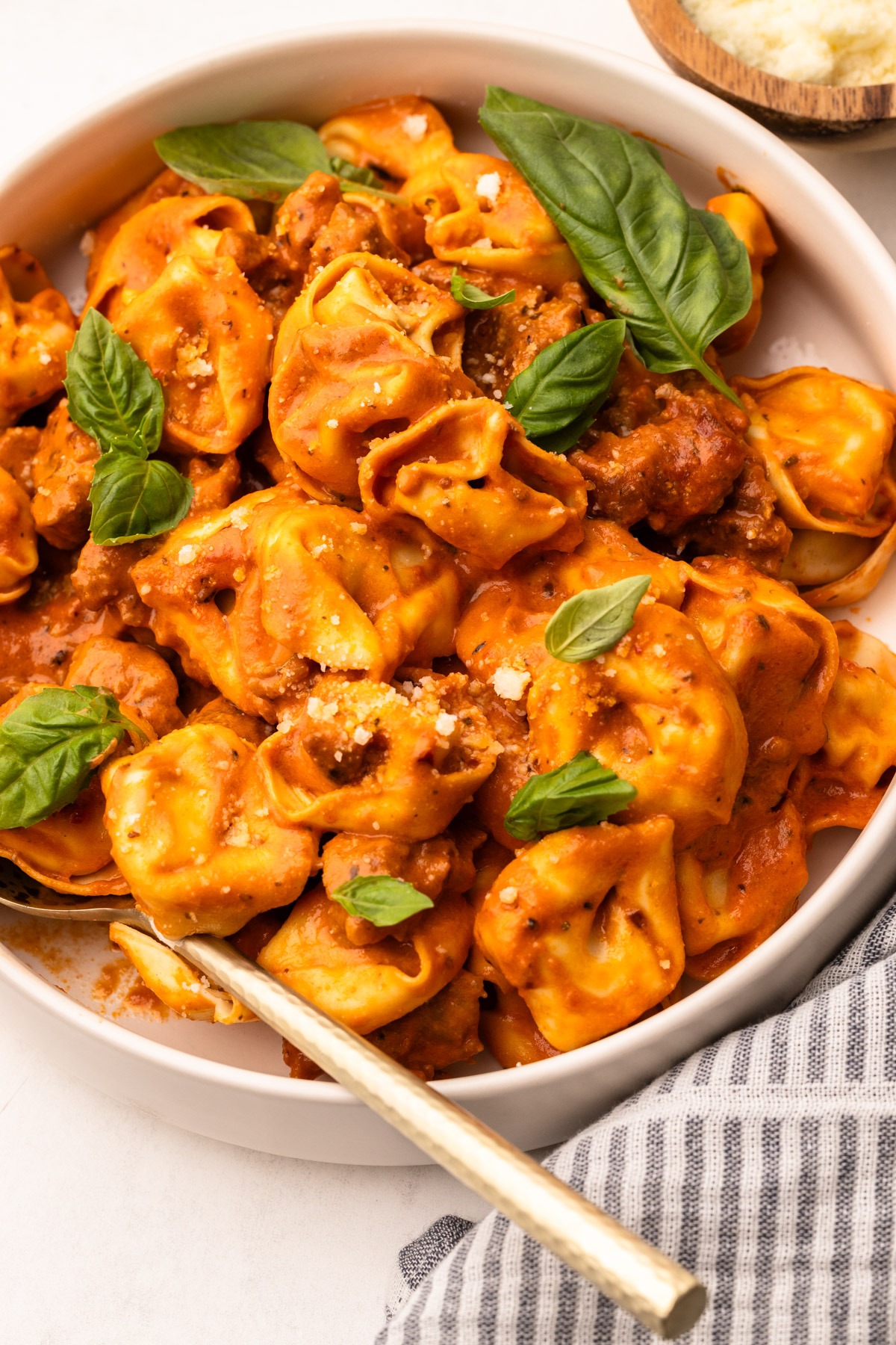 A serving dish of creamy tomato tortellini with sausage, with sprinkles of basil and a spoon.