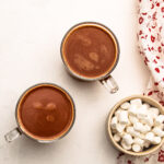 Two mugs of hot chocolate with chocolate chips, with a bowl of mini marshmallows.