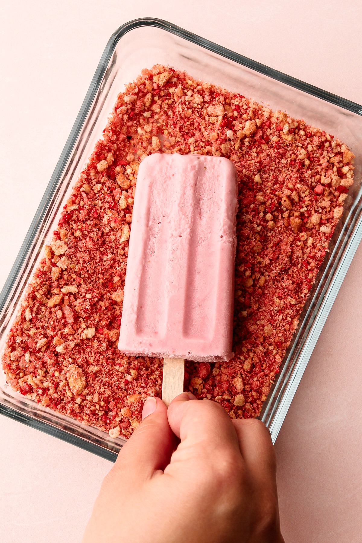 Hand dipping a pink strawberry shortcake popsicle into a crumbly coating mixture.