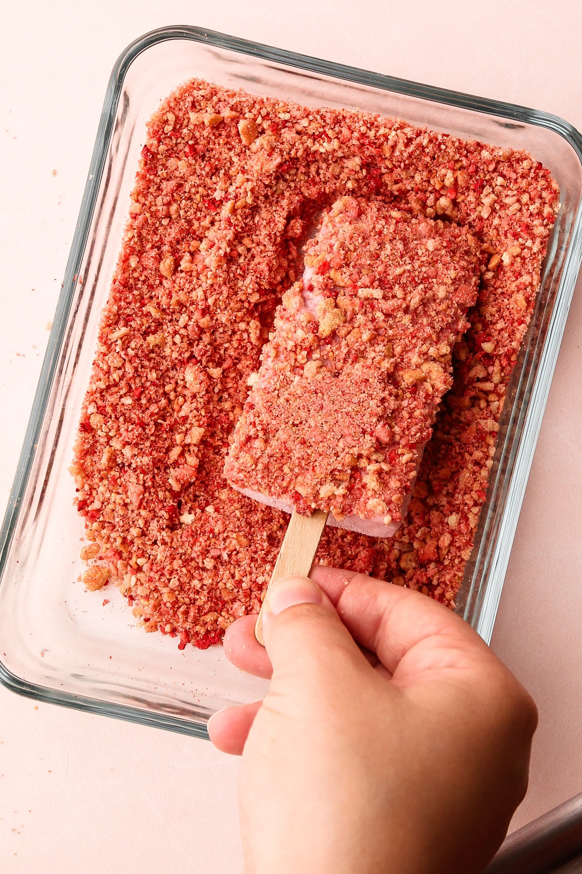 Hand pressing a strawberry shortcake popsicle into a crumbly coating mixture.