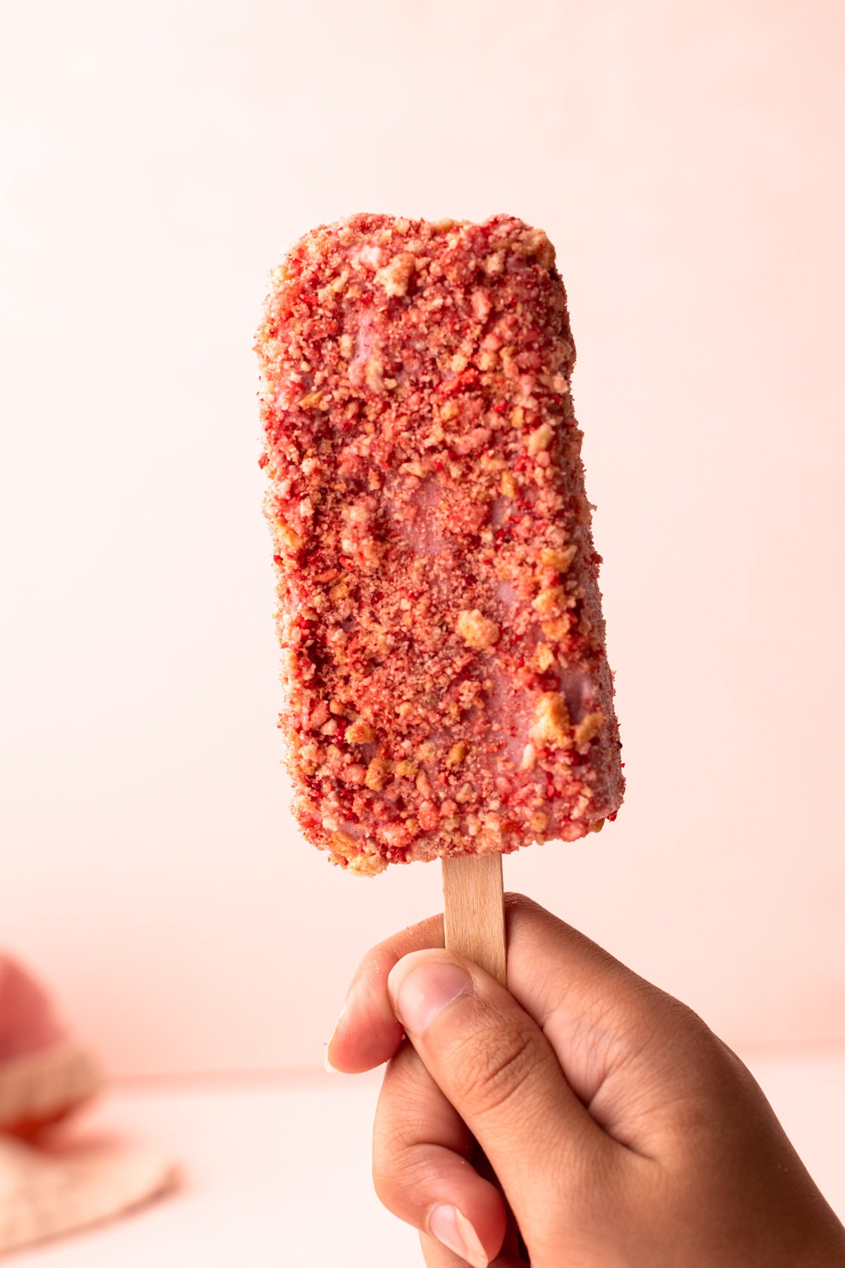 Hand holding a strawberry shortcake popsicle covered in a crumbly coating.