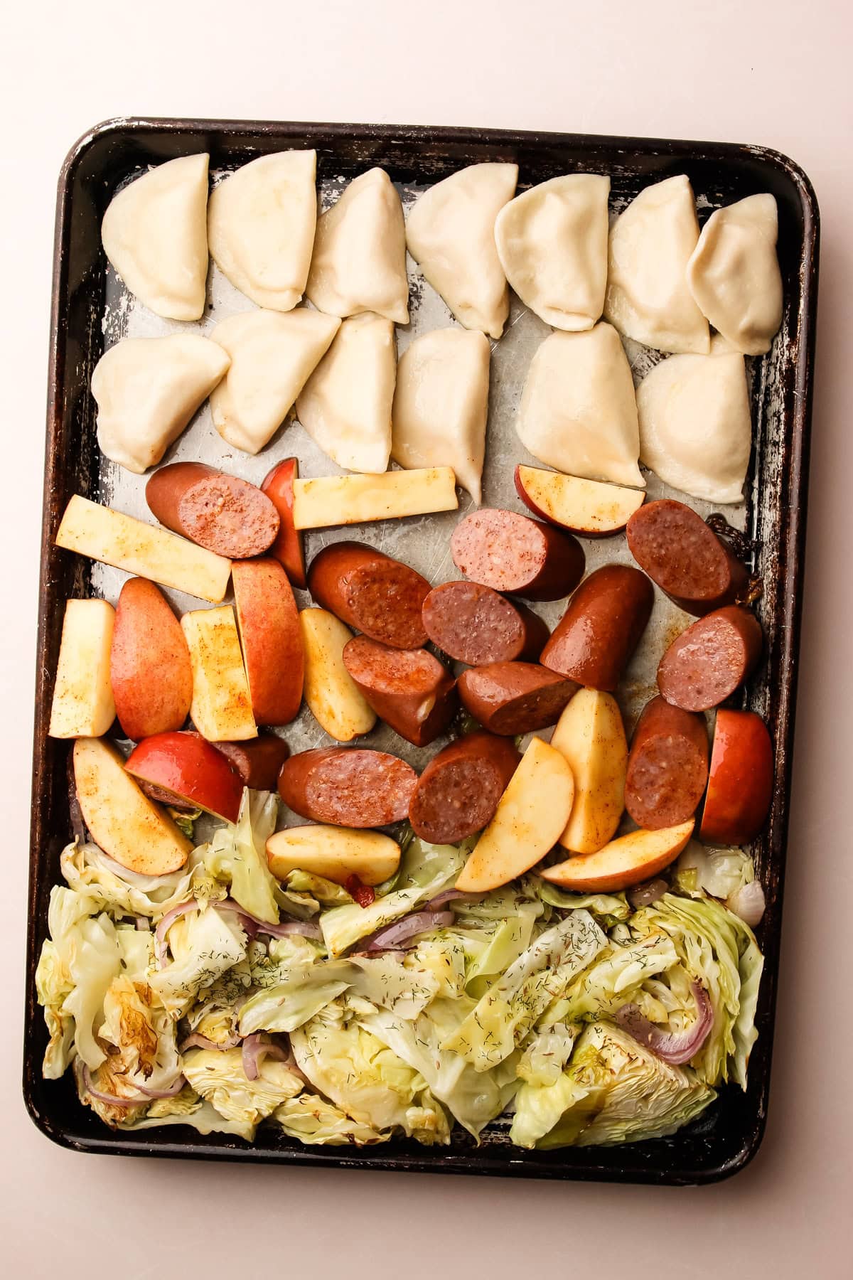 Uncooked pierogis, kielbasa, and sliced apples laid out on a sheet pan with fresh cabbage, prepped for a pierogi and kielbasa recipe.