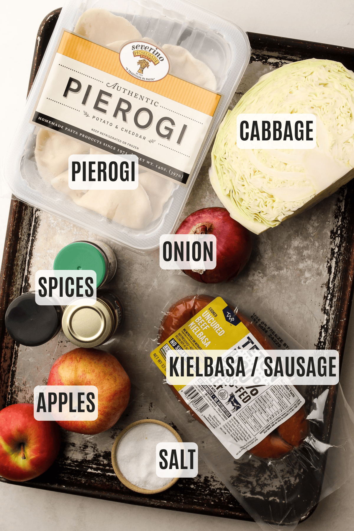 Overhead of all ingredients for this sheetpan pierogi recipe: pierogi, cabbage, onion, apples, kielbasa, spices, and salt, labeled and arranged on a tray.