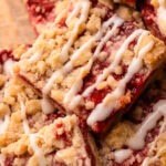 Baked and sliced strawberry crumble bars with a drizzle of icing stacked up on top of one another on a wood board.