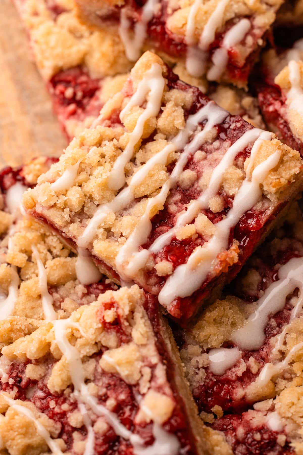 Baked and sliced strawberry crumble bars with a drizzle of icing stacked up on top of one another on a wood board.
