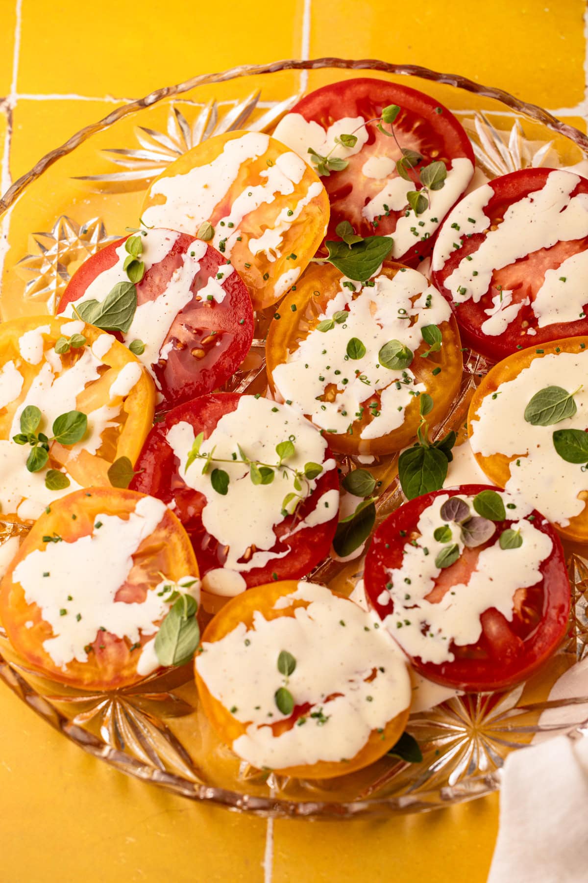 A creamy tomato salad with fresh herbs on a glass plate with a yellow background.
